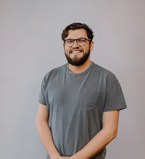 A smiling man with glasses, wearing a gray t-shirt, stands against a plain background.