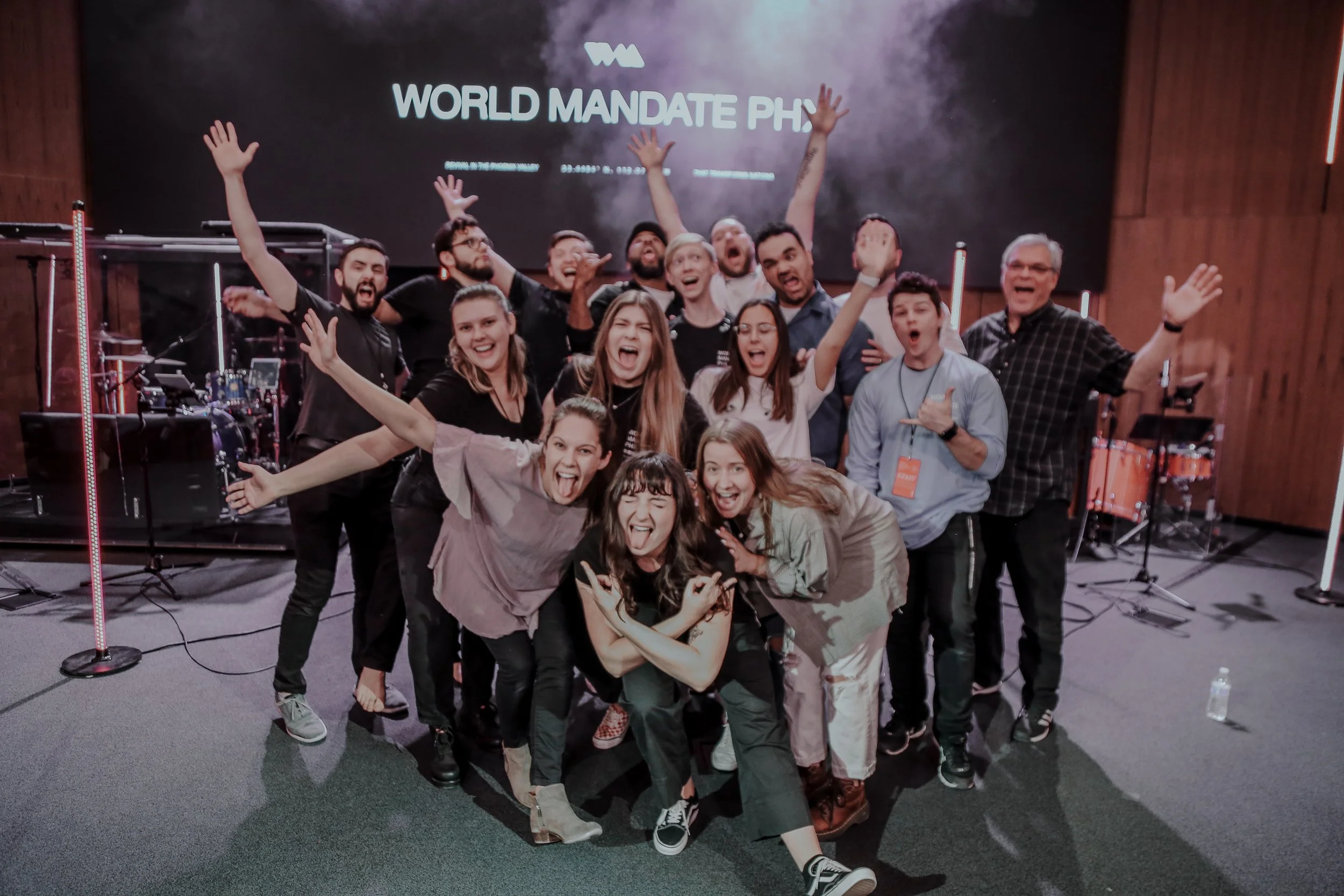 Group of enthusiastic people posing on stage with "World Mandate PHX" sign in the background, some making celebratory gestures.