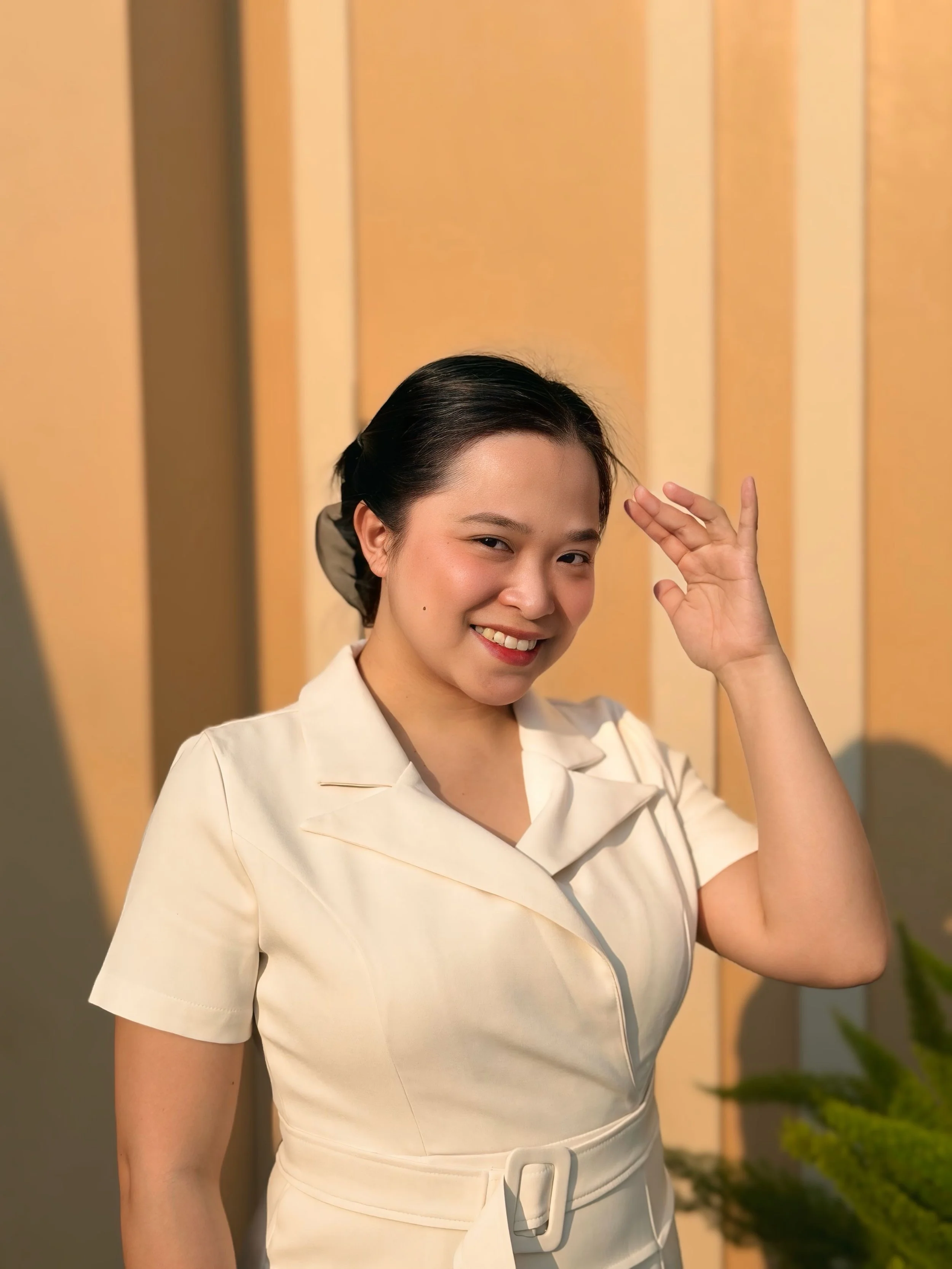 A woman in a cream-colored dress smiling and waving her hand.