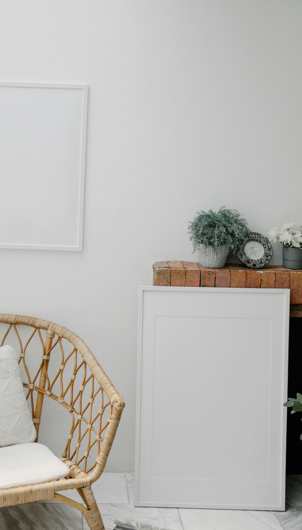 Interior corner with a rattan chair, a white pillow, a framed whiteboard, a brick ledge with potted plants, and a decorative clock.