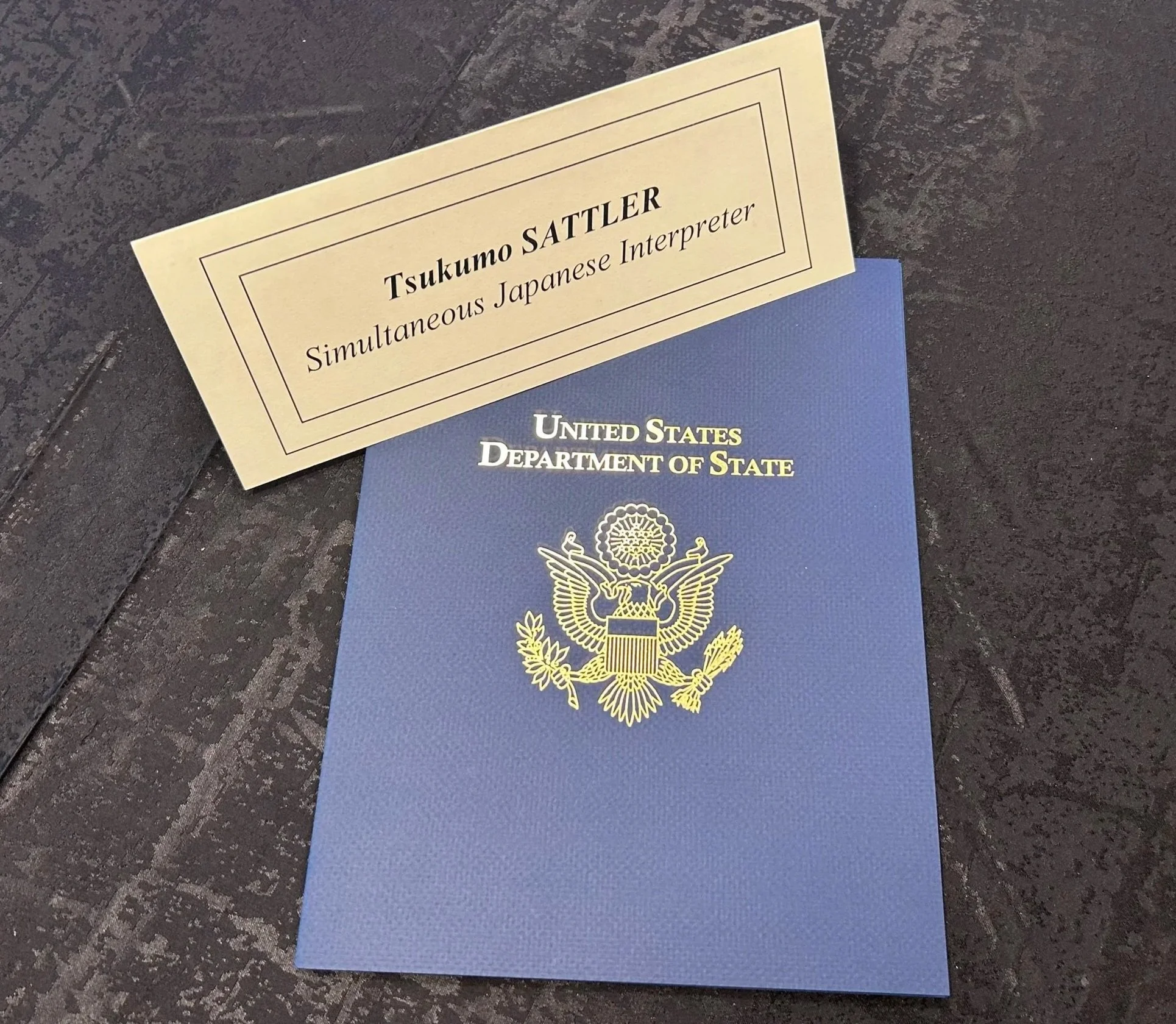 A name placard saying "Tsukumo Sattler, Simultaneous Japanese Interpreter" and a blue portfolio saying "United States Department of State"