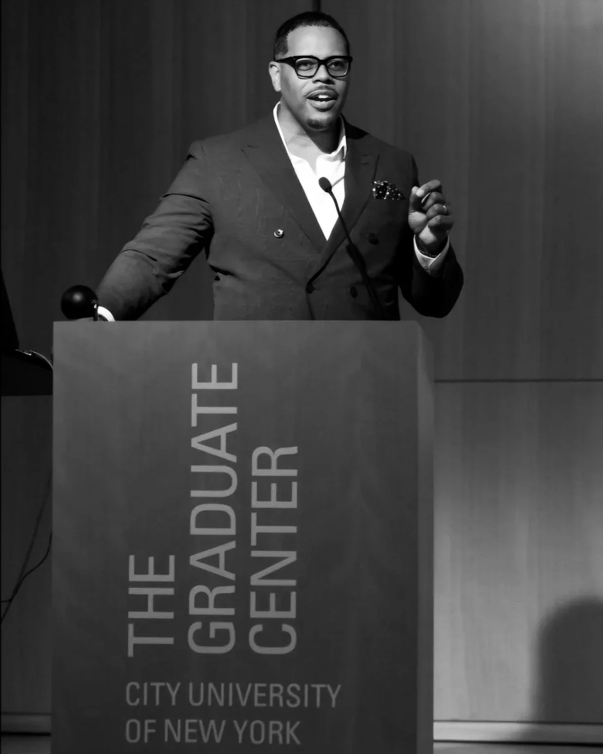 man speaking behind a microphone and podium with the name, "The Graduate Center City University of New York" in black and white