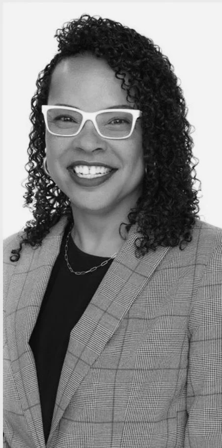 A smiling woman with curly hair wearing white glasses, a plaid blazer, and a chain necklace.