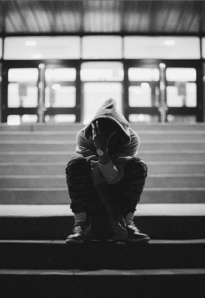 A person wearing a hoodie sitting on stairs with face hidden, in a dark setting with bright windows in the background.