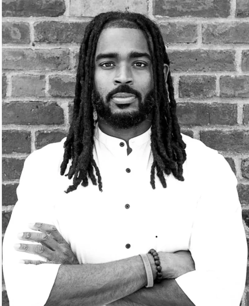 A black and white photo of a man with a beard standing in front of a brick wall, wearing a white shirt with black buttons, with his arms crossed, looking at the camera.