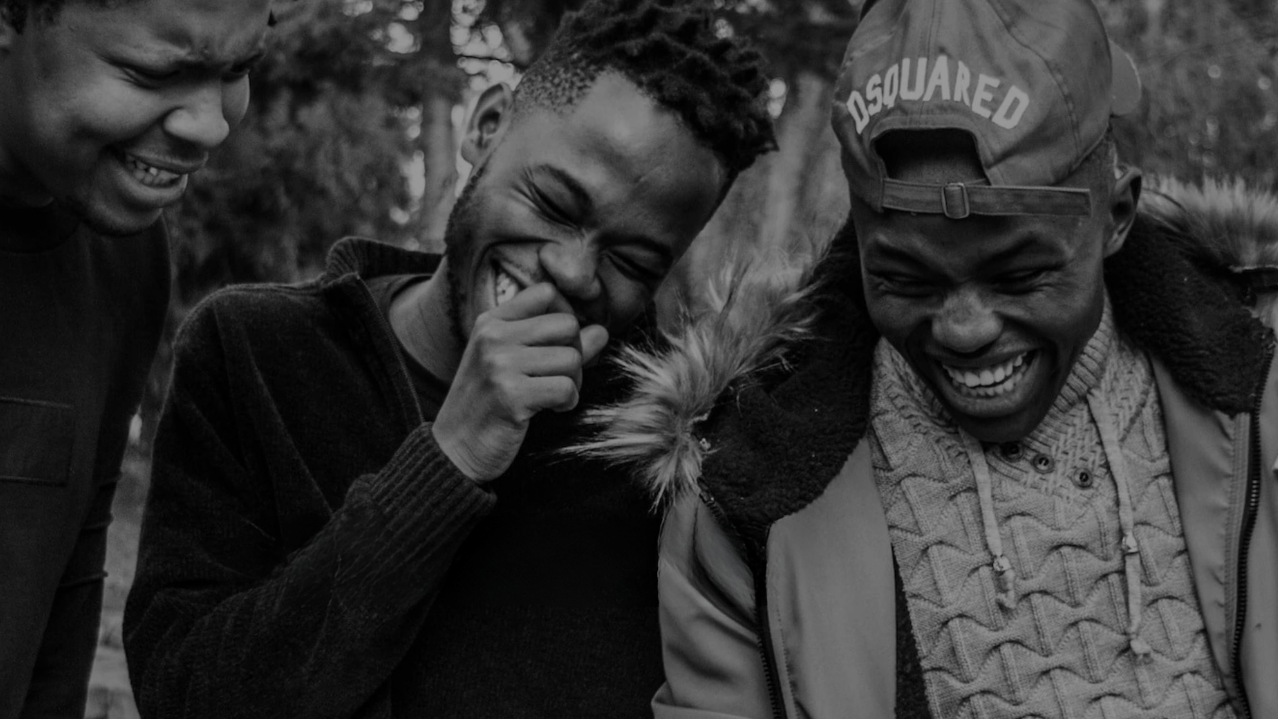 Three people outdoors, smiling and laughing together, one with a hand covering his mouth, wearing a jacket with a fur collar, and wearing a baseball cap with the brand 'Dsquared' on it, with trees in the background.
