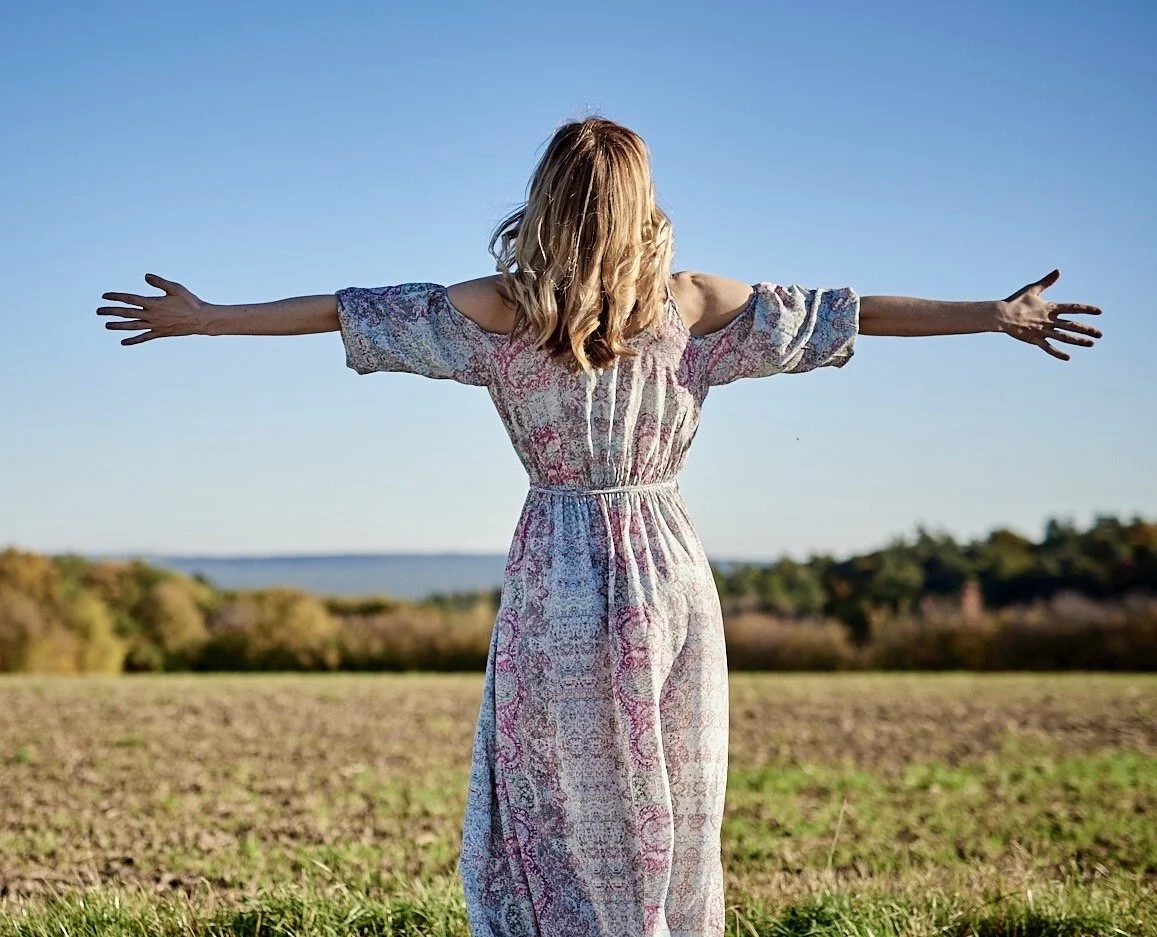 Woman with arms outstretched standing in a field, wearing a floral dress, with trees visible in the background.