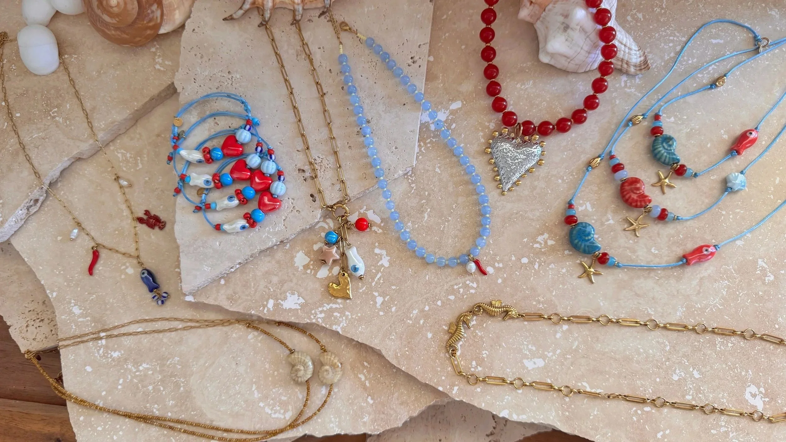 A flat lay of assorted colorful shell and beaded jewelry, including necklaces and bracelets, displayed on light-colored textured stone surfaces.