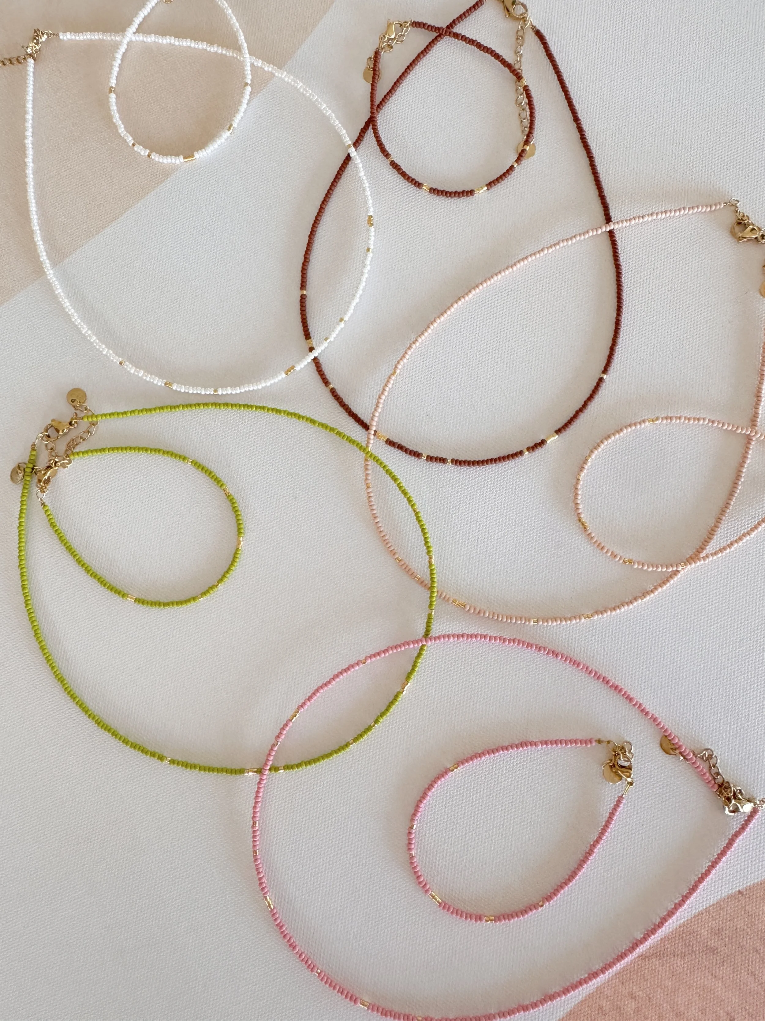 Colorful beaded necklaces in white, red, pink, and green with gold accents, arranged on a fabric surface.