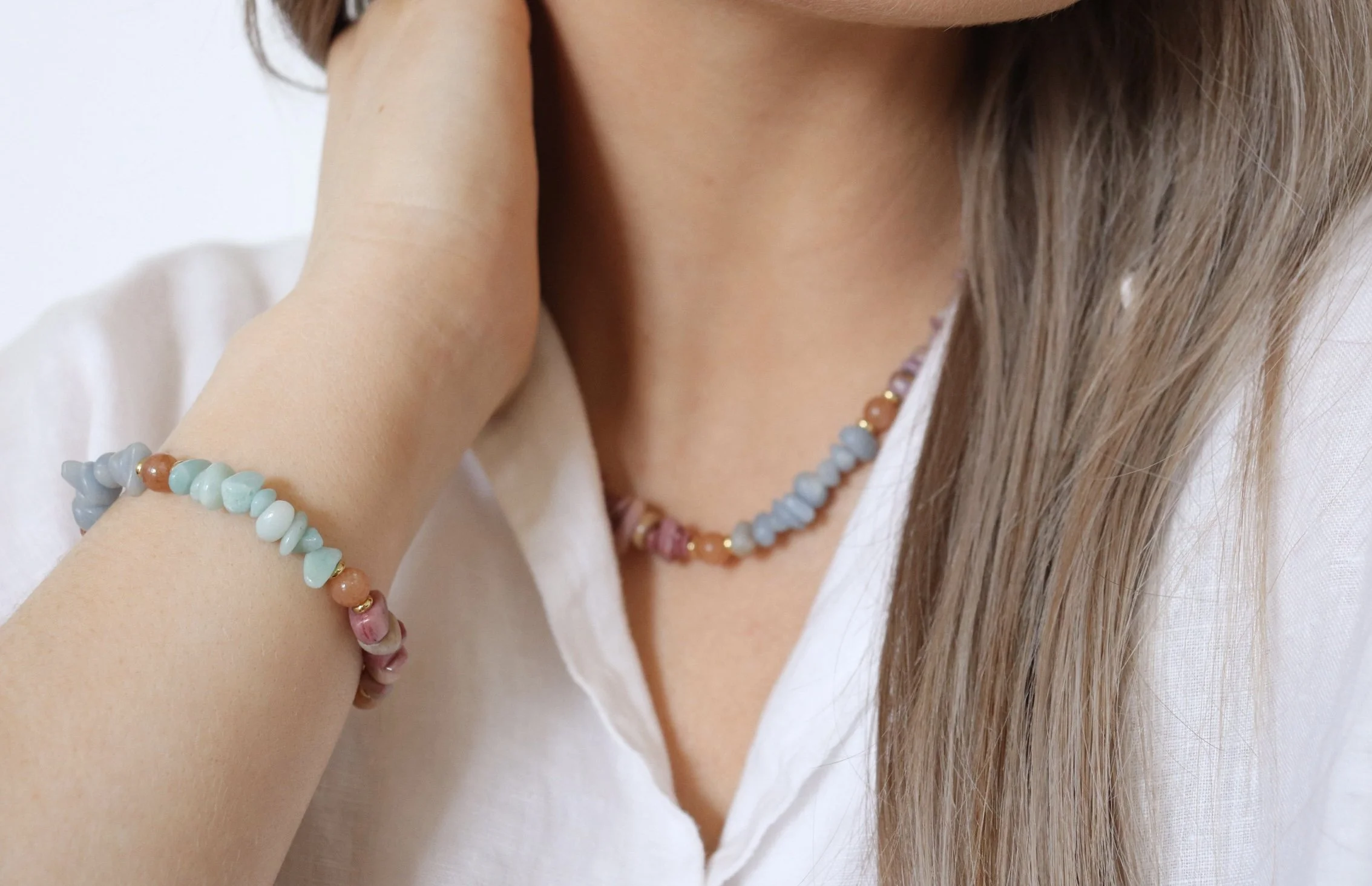 Close-up of a person's neck, shoulder, and upper chest, with a bead necklace and bracelet. The individual has long, light brown hair and is wearing a white top.
