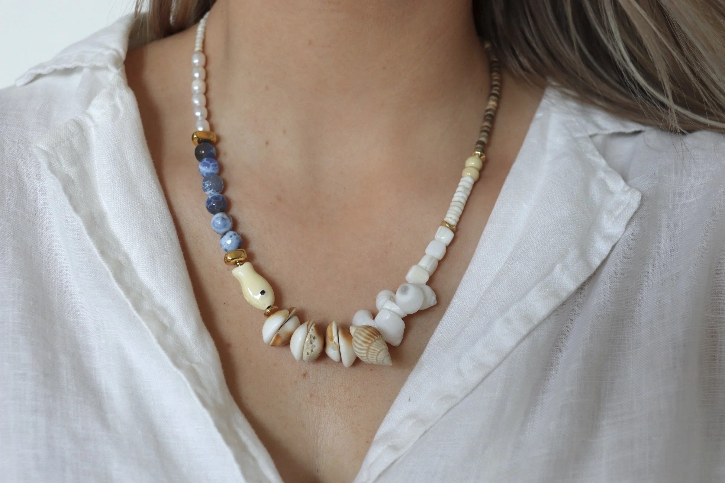 Close-up of a woman's neck and chest wearing layered pearl and shell necklaces over a white, partly unbuttoned shirt.