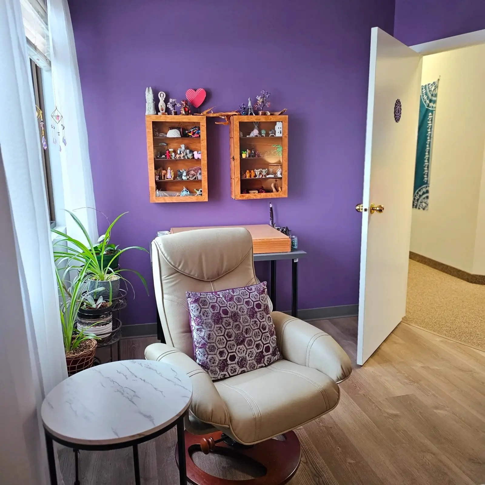 A cozy corner with a beige recliner chair, a purple decorative pillow, a small round white marble side table, potted green plants, and a purple wall with two wooden display shelves containing small decorative items. An open door reveals a hallway wit