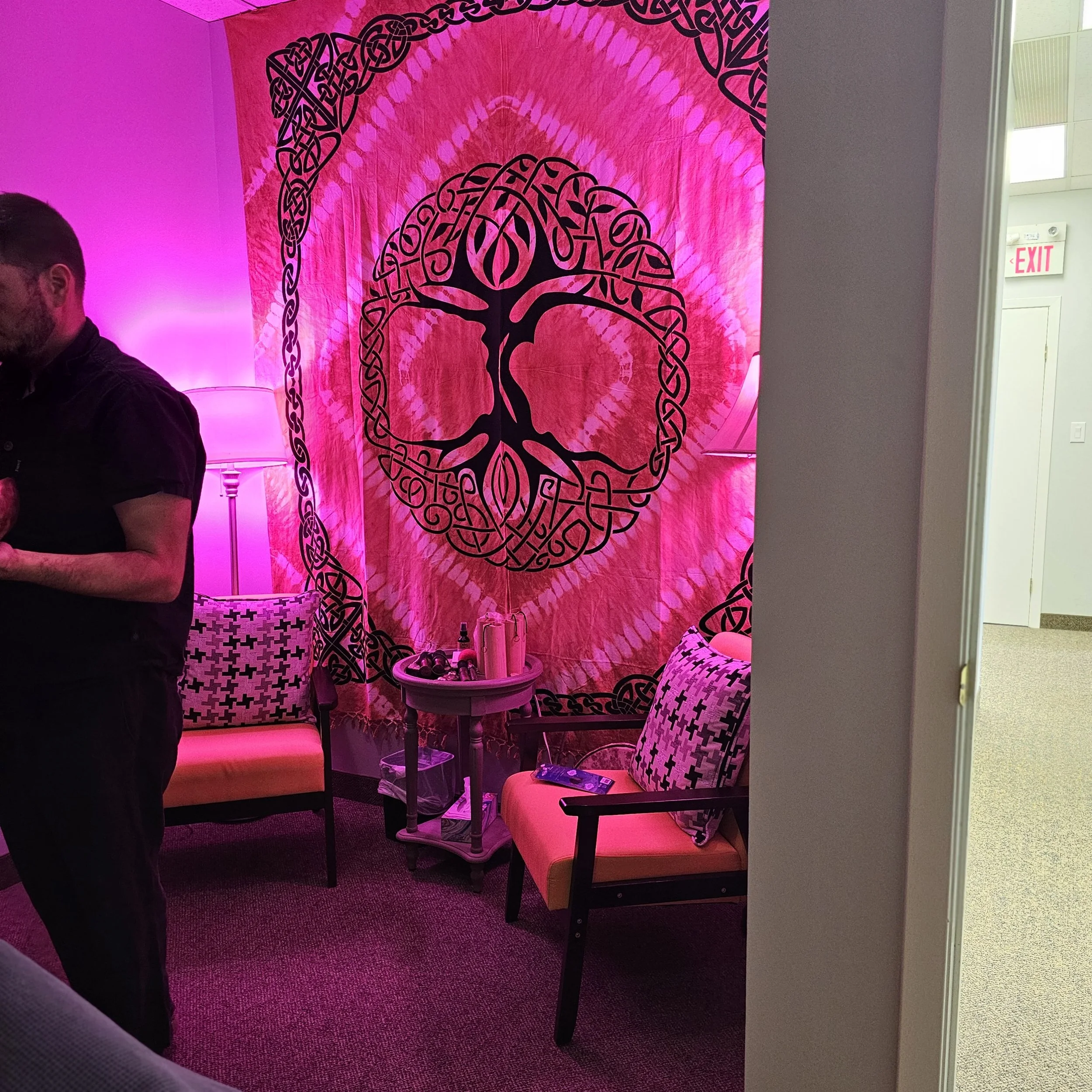 A pink wall with a black Celtic knot style design of a tree. There are four chairs with black and white patterned upholstery, a small round table with drinks, and a pink light illuminating the room.
