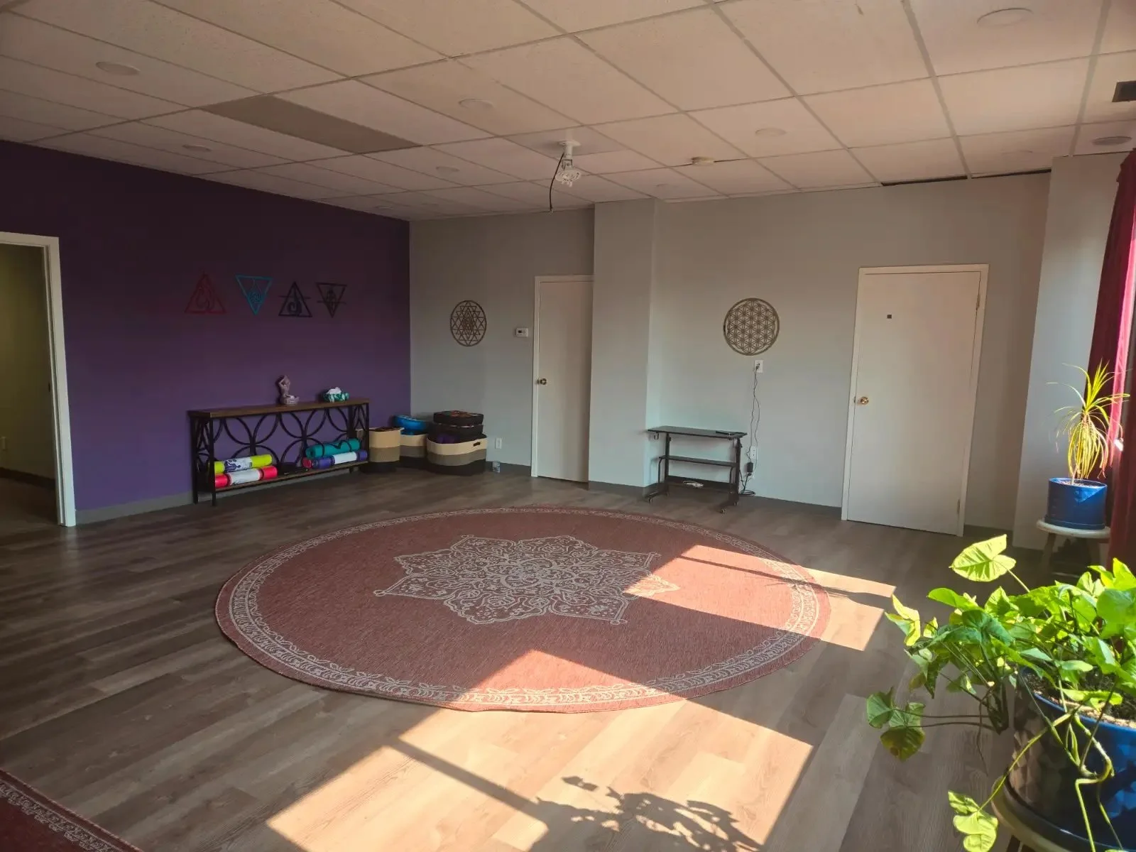 A spacious room with a wooden floor, a large round rug in the center, purple and white walls, decorative wall hangings, a small black shelf against the purple wall with exercise mats, and a potted plant near the window with red curtains.