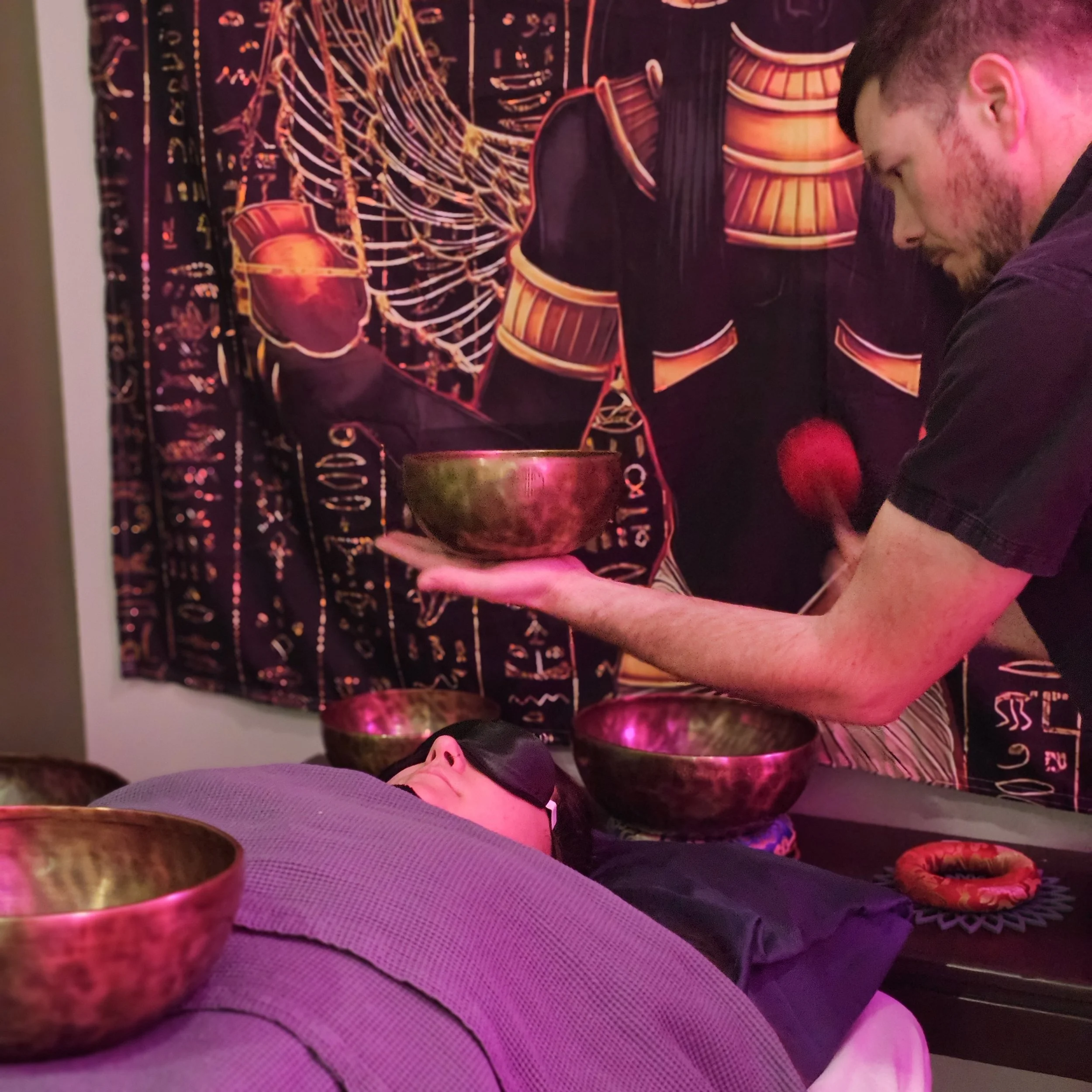 A person with a beard smiling while holding a wooden bowl, with a dark background featuring artwork of drums and vibrant colors.