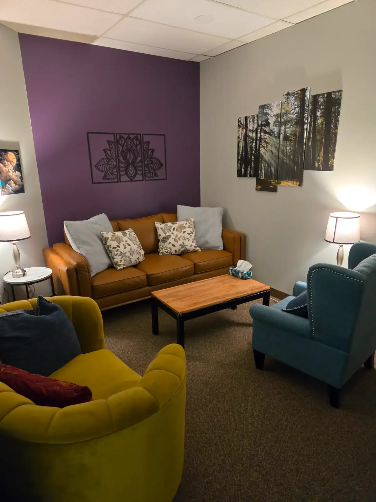 A cozy living room with a yellow armchair, a leather sofa with decorative pillows, a wooden coffee table, blue and green armchairs, and lamps providing warm lighting. Wall art includes a mountain landscape and a forest scene.