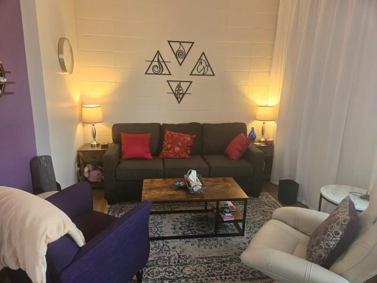 Living room with a black sofa, beige armchair, purple armchair, and wooden coffee table on a patterned rug. Two side tables with lamps, and wall art with geometric designs above the sofa. Various pillows are on the furniture, and there are curtains o
