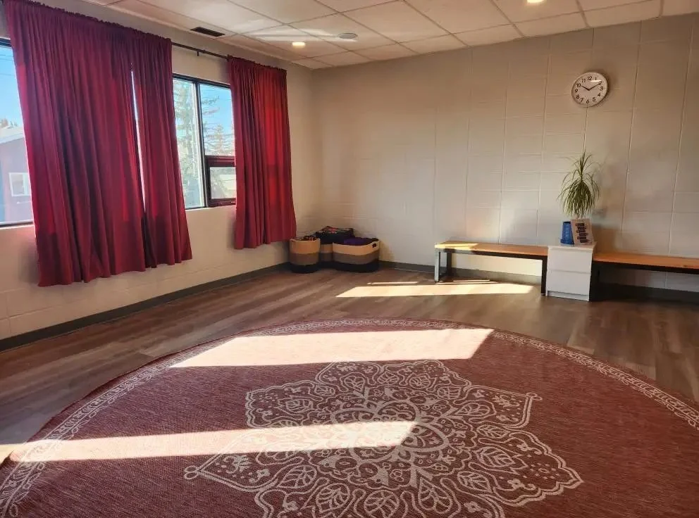 An empty room with large windows, red curtains, a circular rug, and a wooden bench along the wall with a potted plant and a small white cabinet.