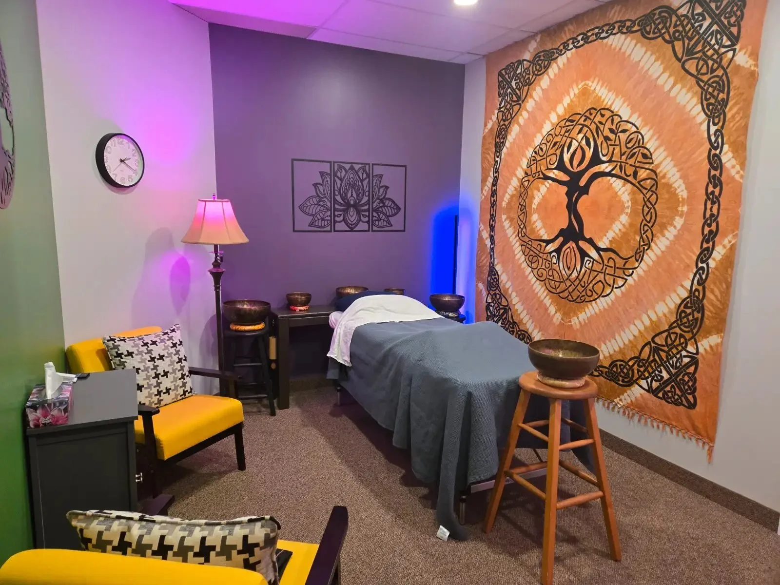 A massage or therapy room with a massage table, a large orange and black tapestry with a tree design on the wall, a wall clock, a floor lamp, chairs with patterned cushions, and bowls on small tables.