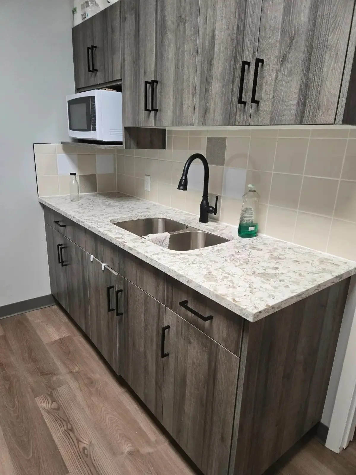 Modern kitchen with a granite countertop, double sink, black faucet, beige tile backsplash, wood cabinets with black handles, microwave, and cleaning supplies on the counter.