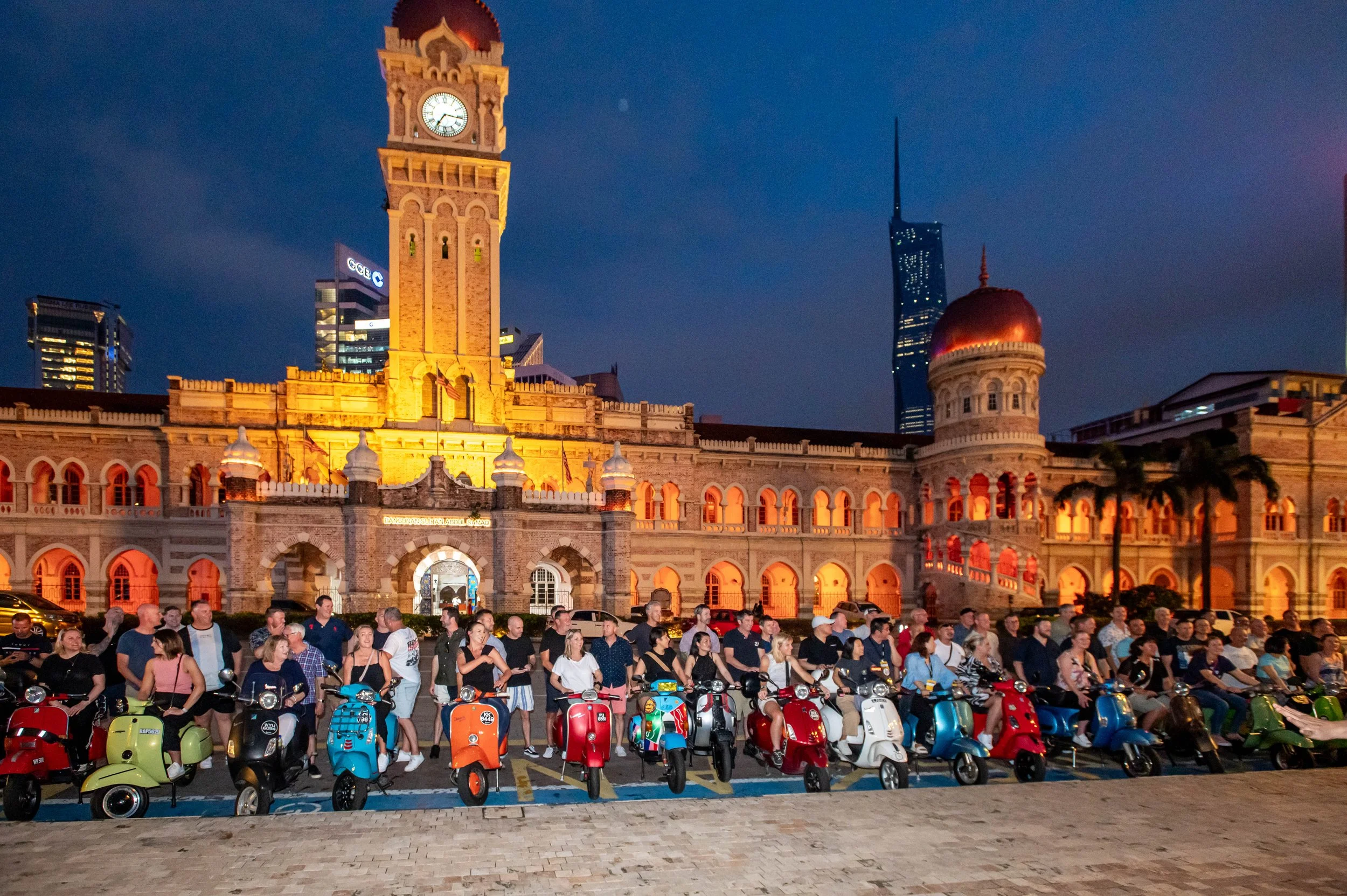 Group of people gathered around colorful scooters in front of a historic building with a clock tower at night.