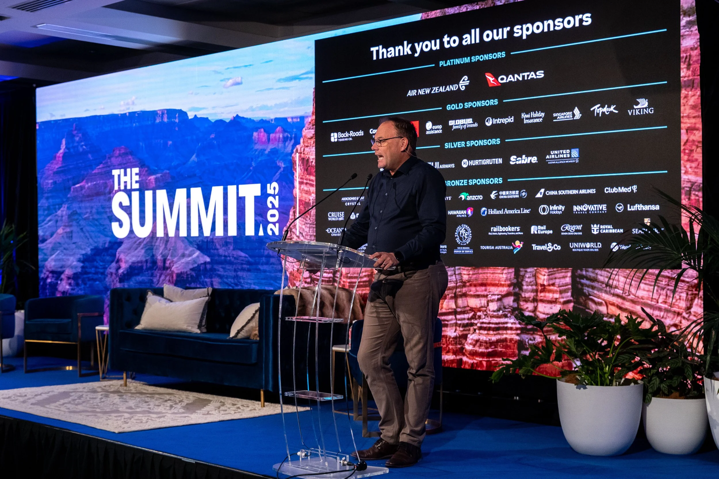 A man speaking at a conference stage with a large screen behind him displaying sponsors and the event name 'The Summit 2023'. The stage has blue carpeting, a glass podium, a black couch with cushions, and potted plants.