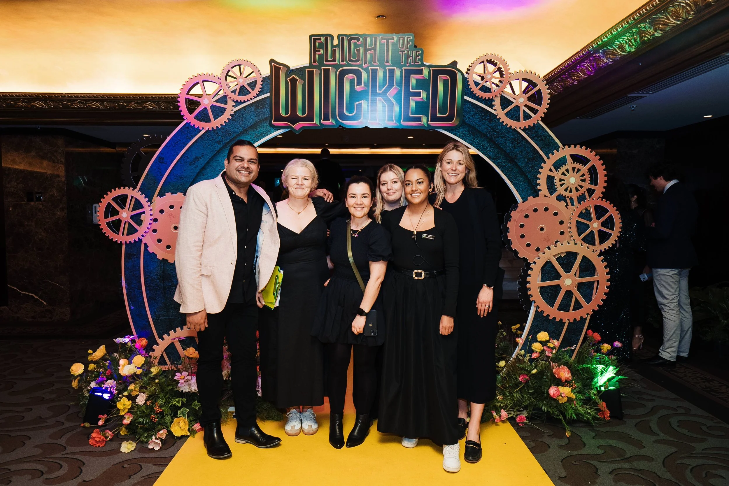 Group of six people smiling and posing in front of a decorative arch with the text 'Flight of the Wicked' at an indoor event.