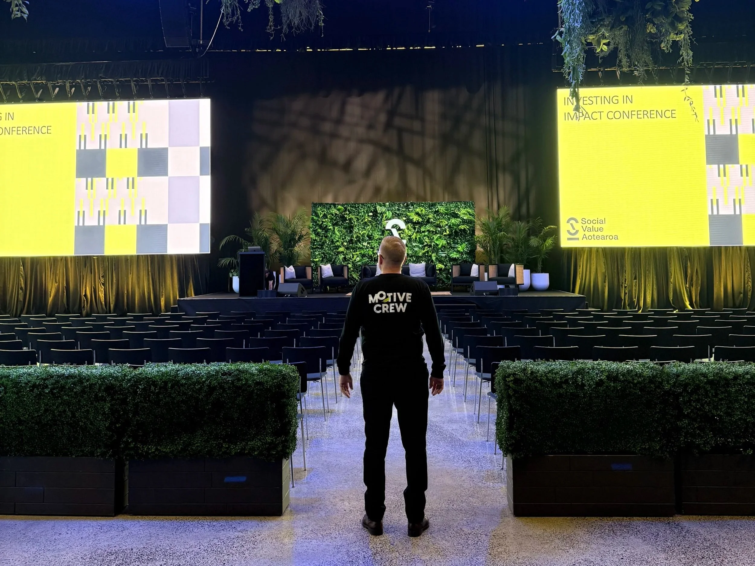 An empty conference hall with rows of chairs facing a stage with large two yellow screens displaying the words "Investing in Impact Conference" and the logo of Social Value Aotearoa. A person wearing a black hoodie with the words "Motiv Crew" on the back is standing centrally, facing the stage, in front of two large potted plants and several chairs.