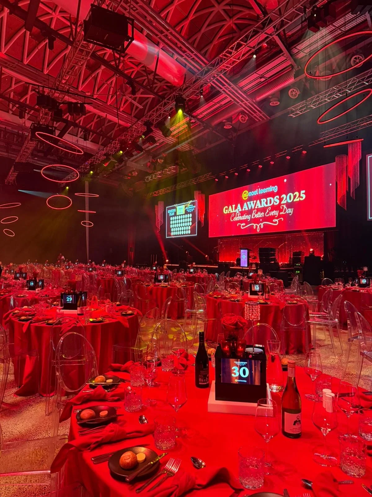 Ballroom decorated for a gala event with round tables covered in red tablecloths, transparent chairs, and place settings. A large screen displays 'GALA AWARDS 2025,' with additional seating and stage lighting in the background.