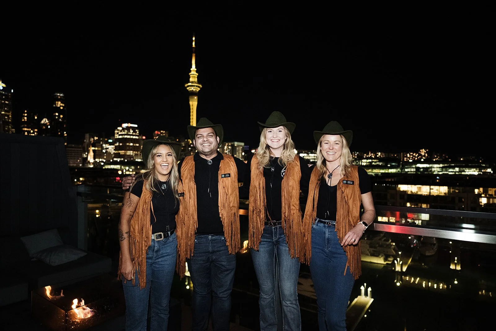 Four people dressed in cowboy costumes standing on a rooftop at night with the city skyline and a tall tower in the background, smiling for the camera.
