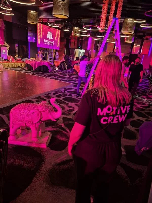 A woman with pink hair and a black shirt that says 'MOTIVE CREW' is standing in a decorated event space with pink lighting. There is a pink-looking elephant statue in the foreground and pink and gold decorations, including hanging pink balls, on the 