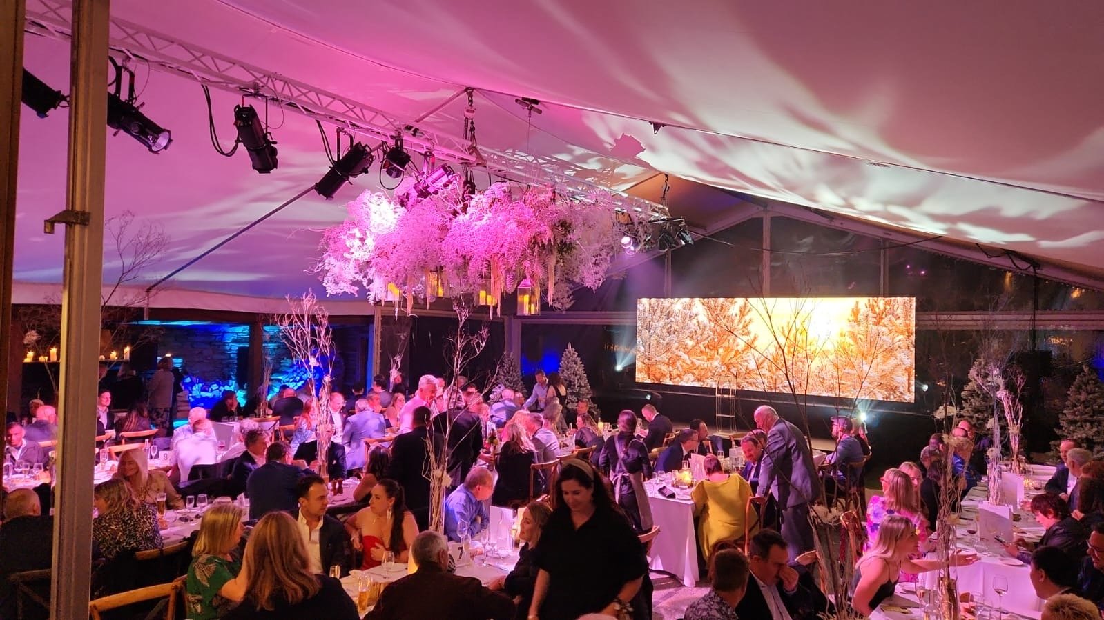 A large event tent filled with people seated at decorated tables. The tent is adorned with pink and white floral decorations, trees, and hanging lanterns. There is a large LED screen displaying autumn-themed imagery and stage lights illuminating the scene.