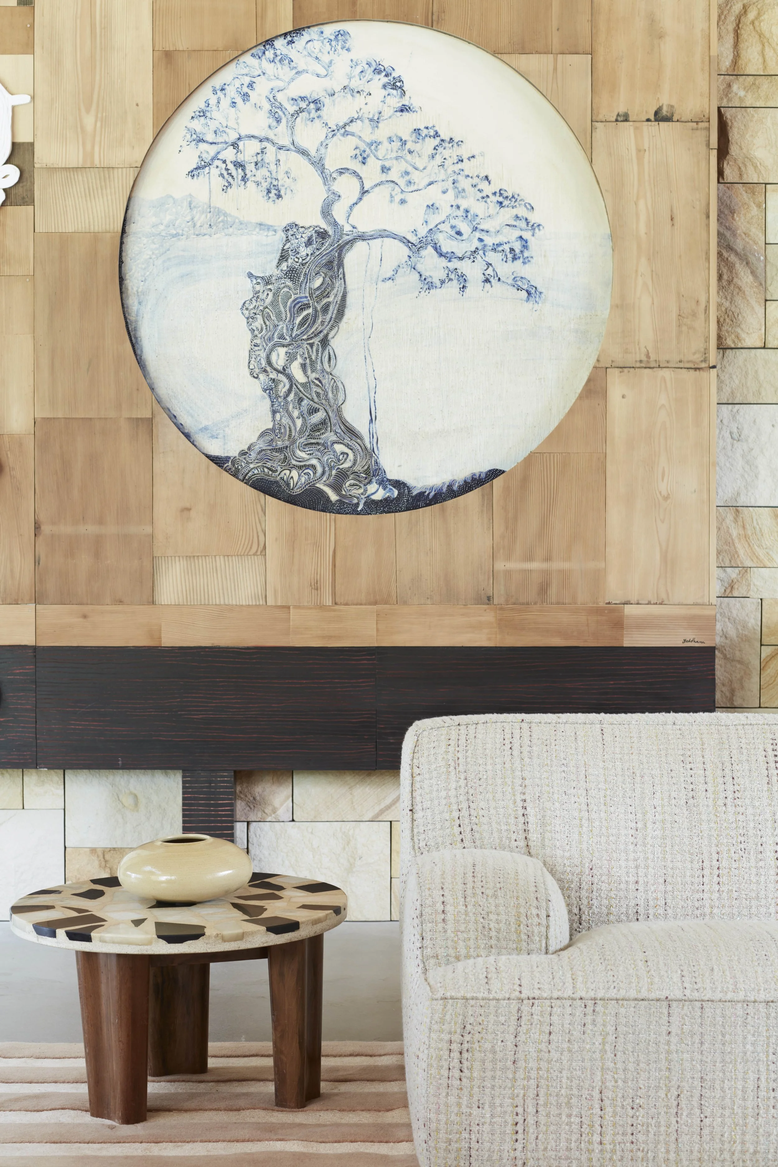 Living room with a round wall art featuring a blue tree, a small round table with a black-and-white geometric pattern and a beige ceramic vase, and a cream-colored upholstered armchair with a patterned fabric, set against a wooden wall.