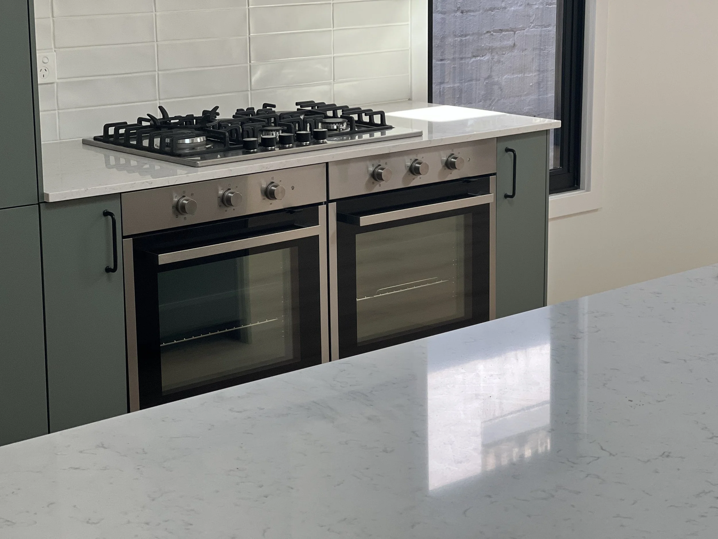 A modern kitchen stove with four burners and an oven beneath, built into green cabinetry with black handles. The kitchen features a white marble countertop and a backsplash with white subway tiles. There is a window with a black frame beside the stov