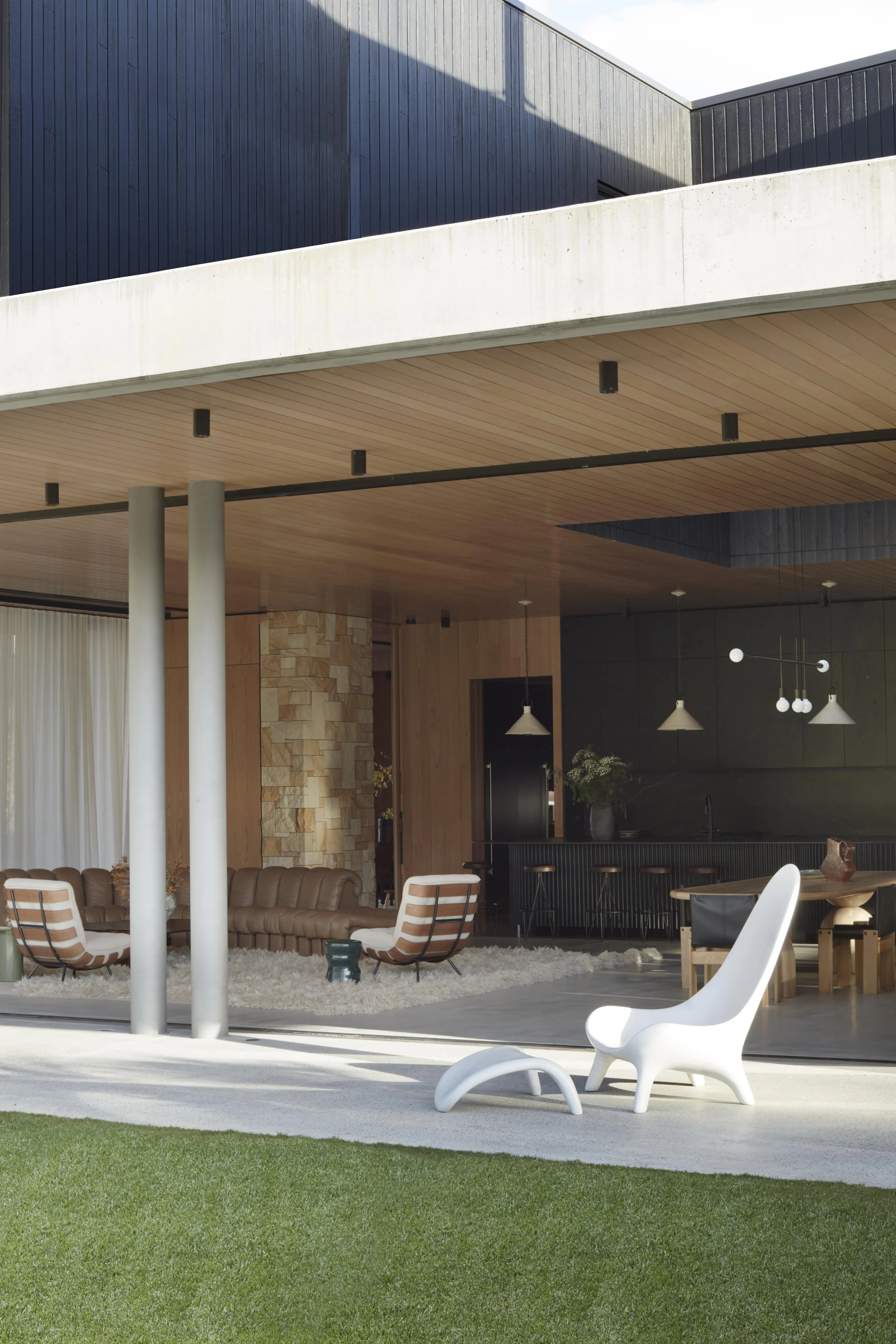 Modern house with outdoor living area, including white and brown chairs, a brown leather couch, a white lounge chair, a rug, and a dining table with chairs, all under a covered patio with pendant lights, with a green lawn in the foreground.