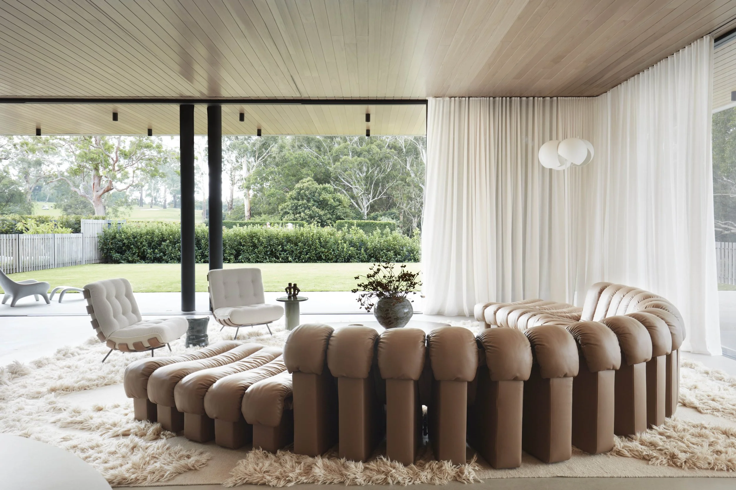 Modern living room with beige tufted sofa, white armchairs, shaggy rug, and floor-to-ceiling windows with white curtains overlooking greenery.
