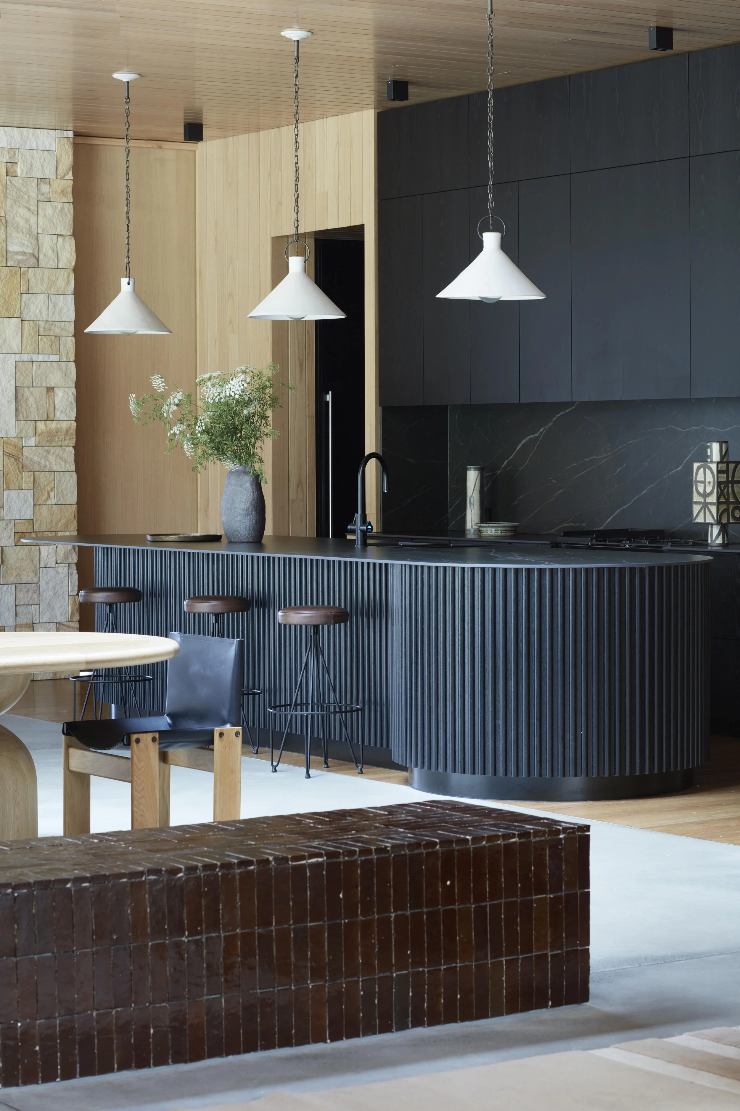 Modern open kitchen with black and wood cabinetry, a black marble backsplash, a curved black ribbed kitchen island, pendant lights, and decorative plants.
