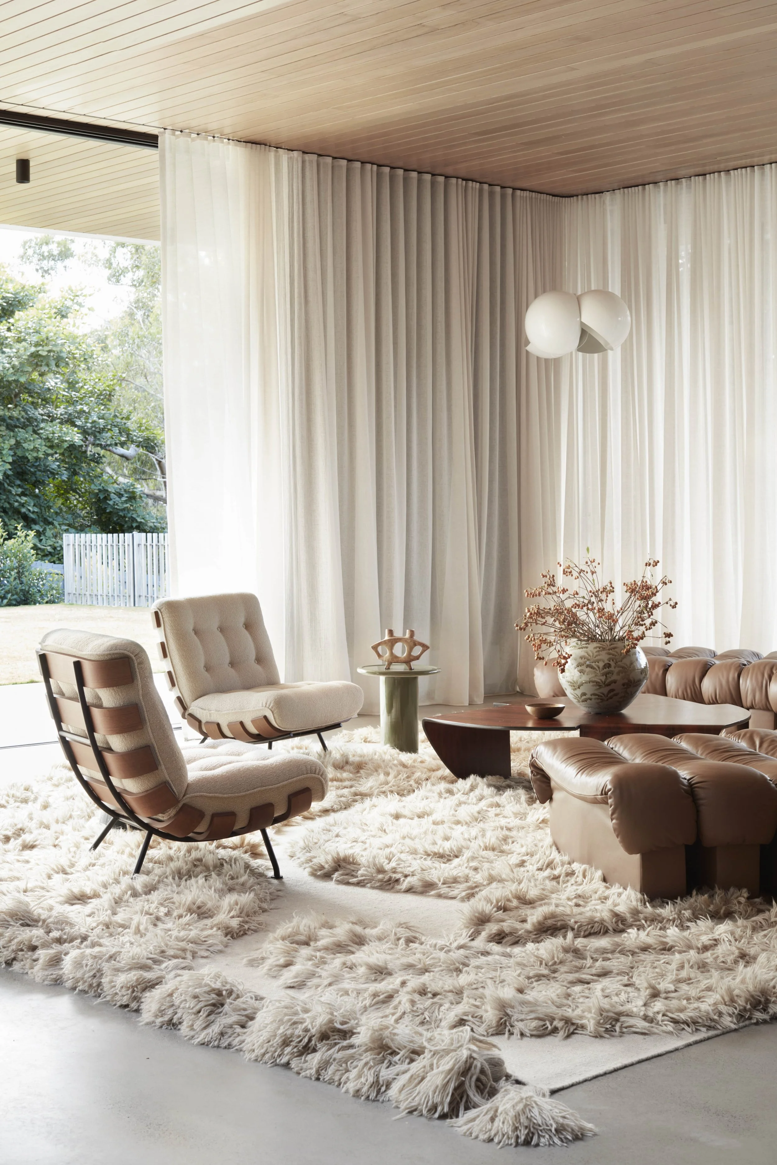 A living room with beige armchairs, a large shaggy white rug, a wooden coffee table, a large decorative vase with dried flowers, and a modern pendant light hanging from a wood-paneled ceiling. Floor-to-ceiling white curtains cover large windows revea