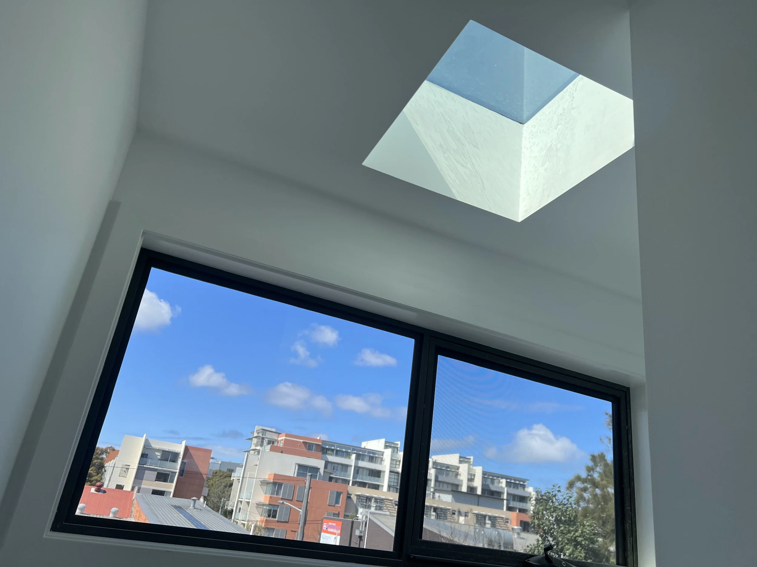 Photo taken from inside a building showing a ceiling skylight and large windows with a view of a cityscape with modern residential buildings and a blue sky with some clouds.