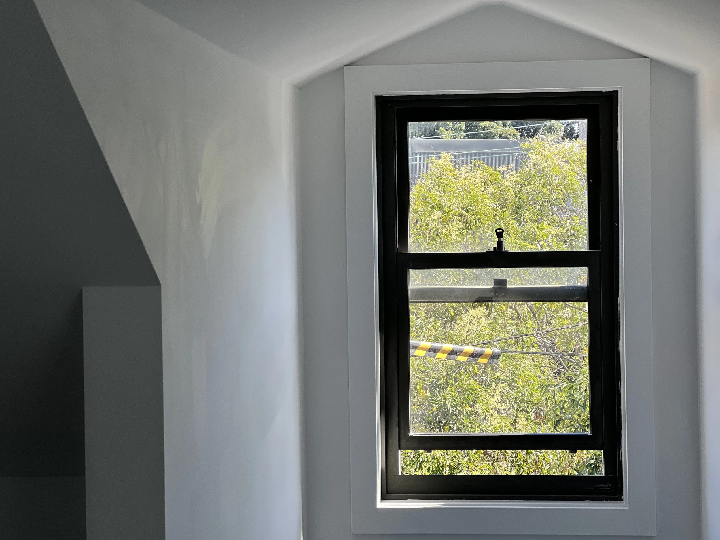 A window with a black frame in a white wall, showing green trees outside and a yellow and black striped barrier at the bottom.