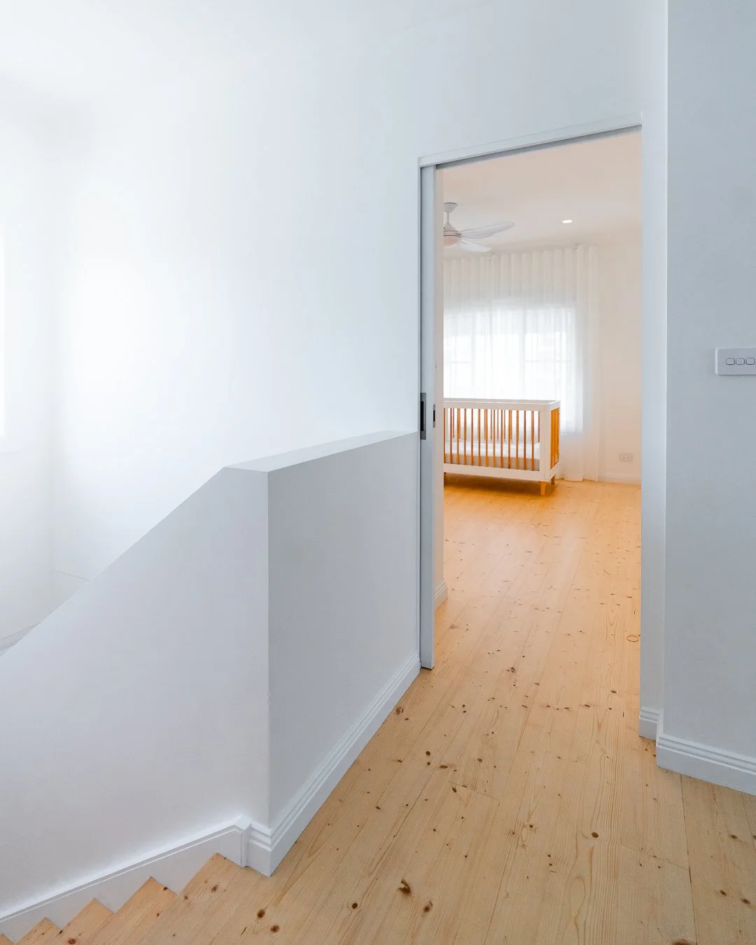 View of an empty room with light wooden flooring and white walls, through an open door from a hallway. A crib is visible in the adjacent room with sheer white curtains and a ceiling fan.