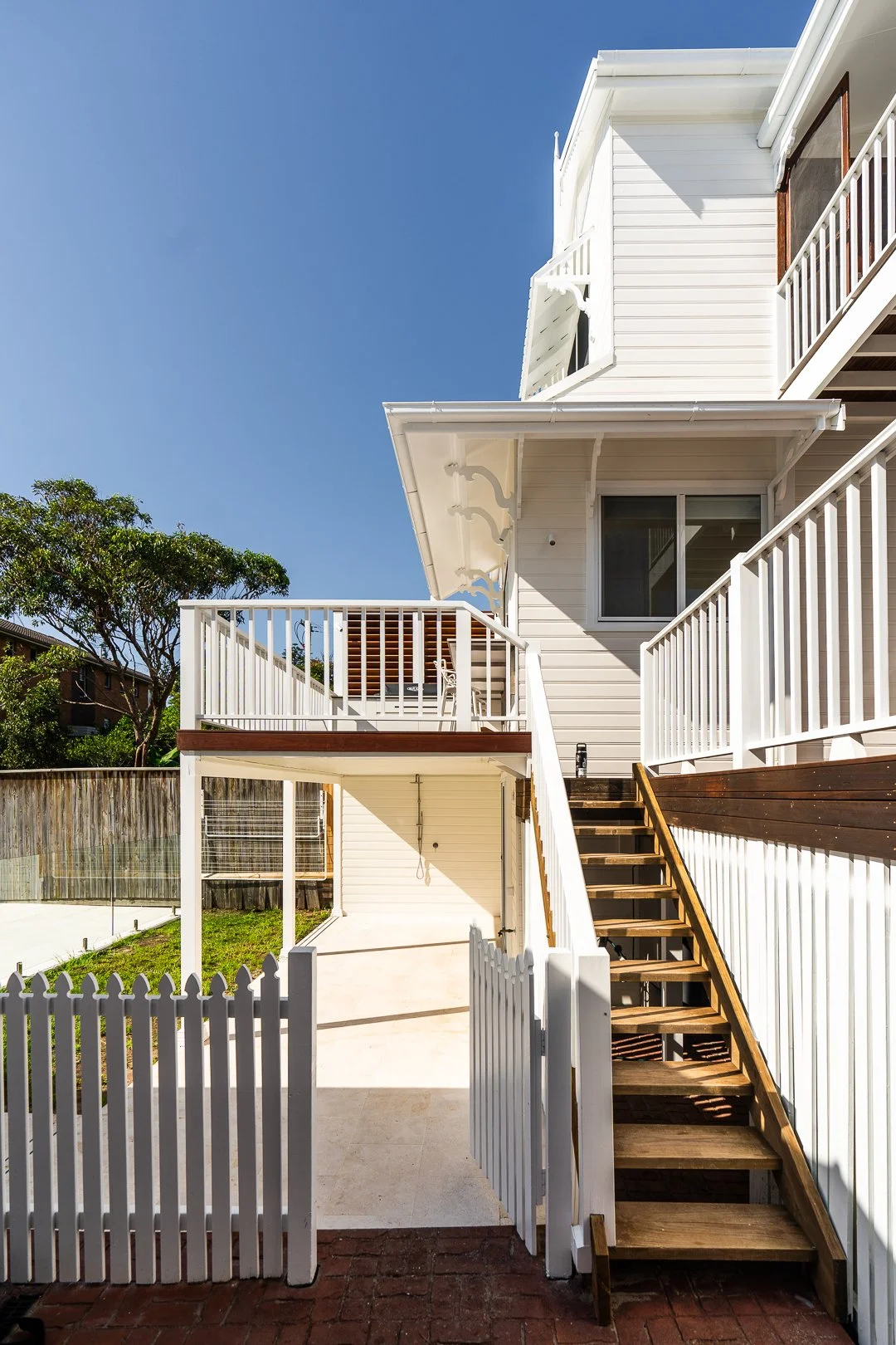 A multi-story white house with wooden stairs leading to a deck, fenced yard with a grass patch, and a clear blue sky.
