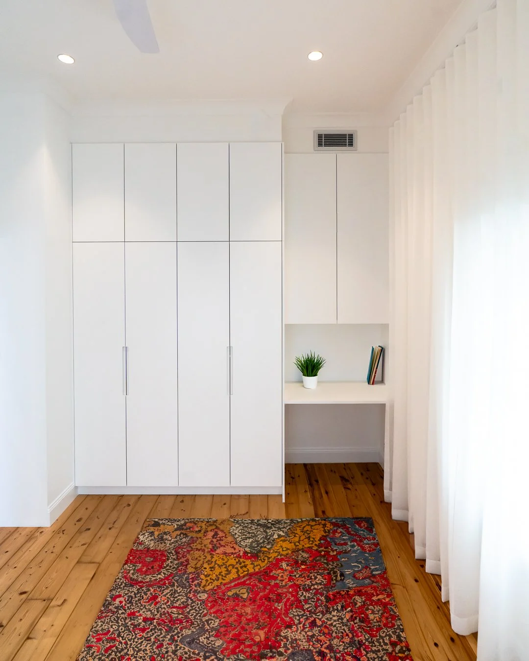 Bright room with white built-in cabinets, a colorful patterned rug, wooden floor, a small shelf with books and a green plant, and sheer white curtains.