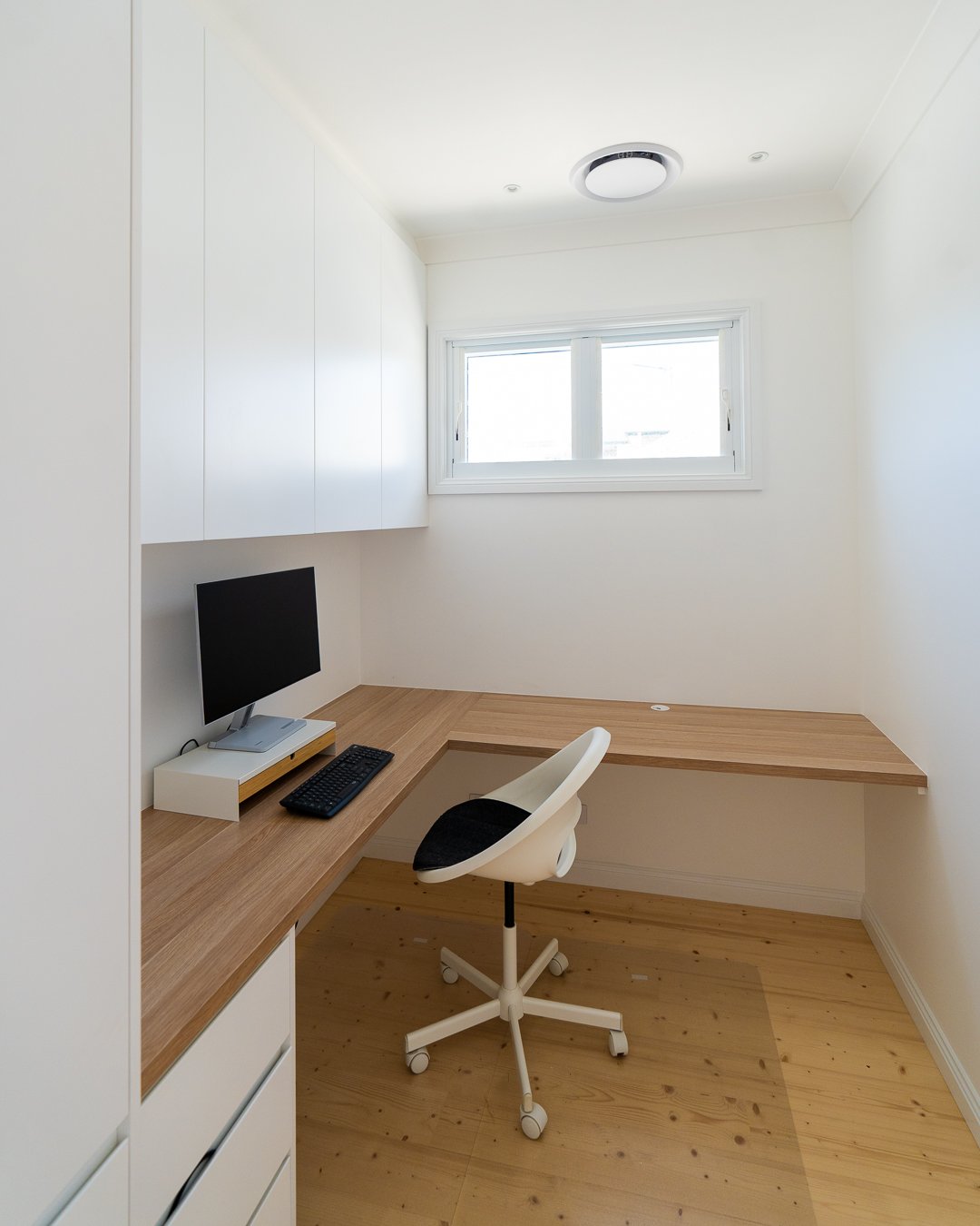 Empty home office with a wooden desk, white chair, black computer monitor, and a window with natural light.