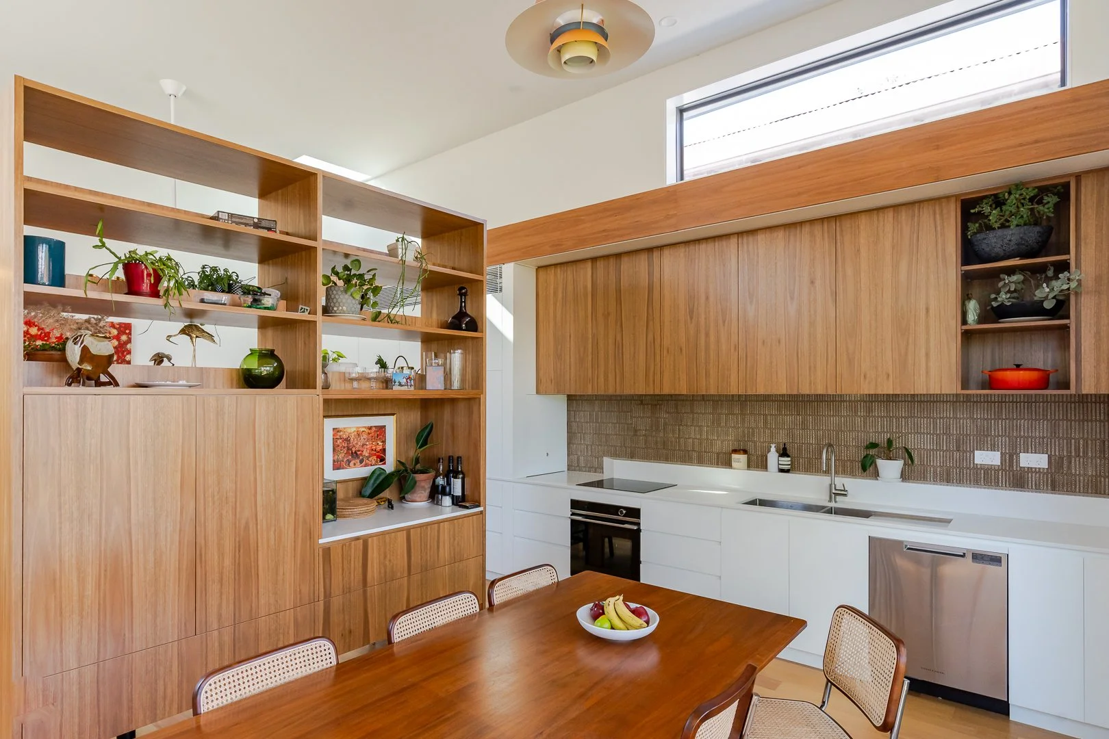 Modern kitchen with wooden cabinets, open shelving filled with plants and decorative items, white countertops, brick backsplash, and a wooden dining table with a fruit bowl.