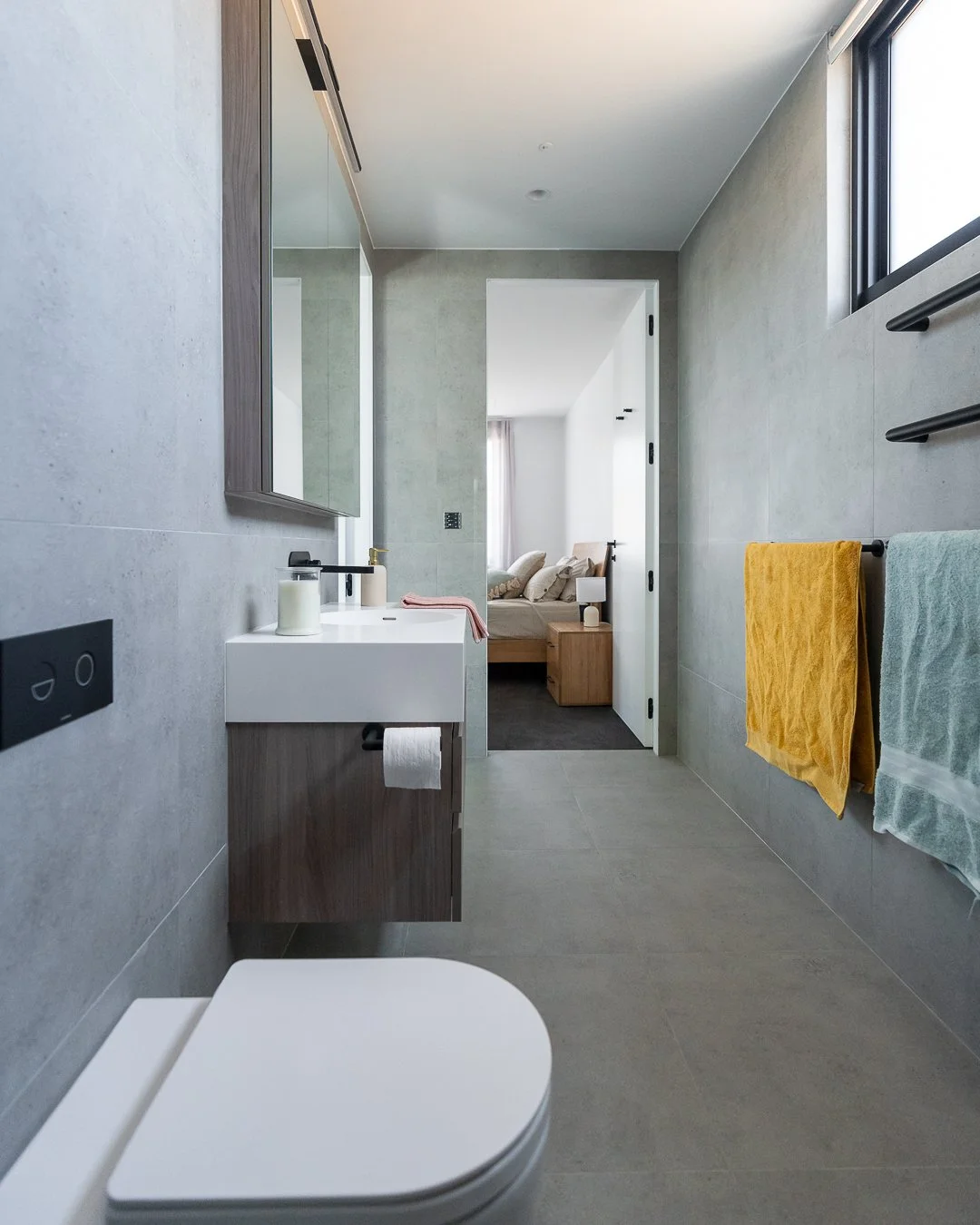 Modern bathroom with a white vanity, large mirror, gray tiled walls, towel rack with yellow and green towels, and a window. Through the doorway, a bedroom with a bed, nightstand, and lamp is visible.