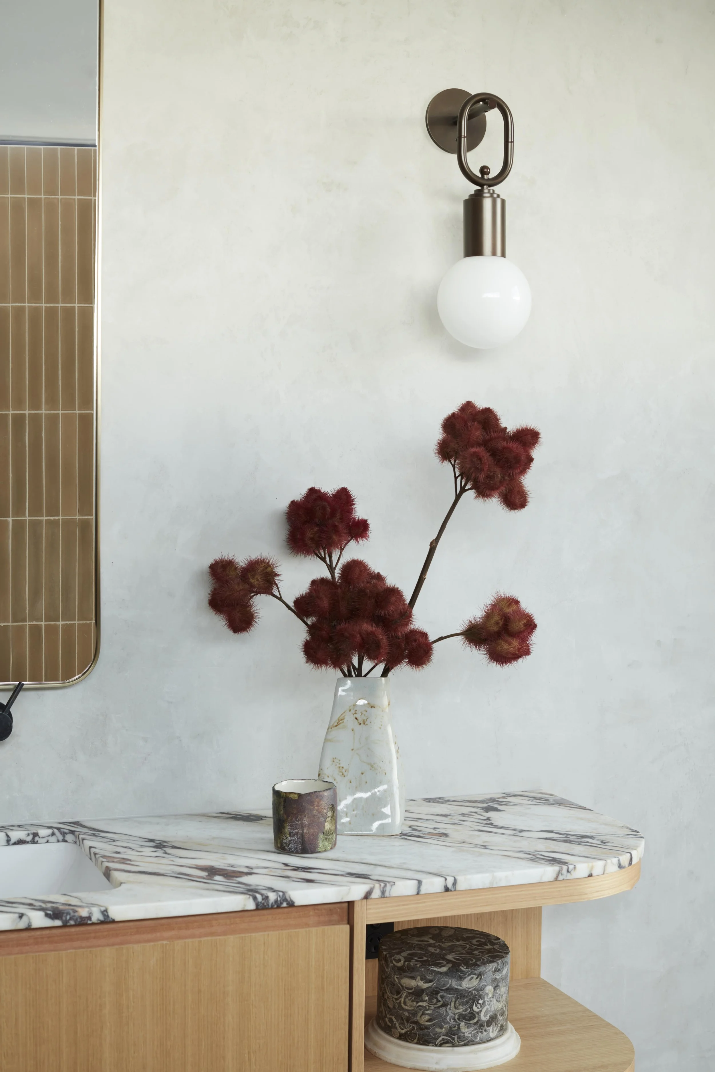 A white marble countertop with a wooden base, decorated with a tall white vase holding red dried flowers, a smaller dark ceramic pot, and a marble-patterned cylindrical container. A modern wall sconce with a frosted glass globe is mounted on the whit