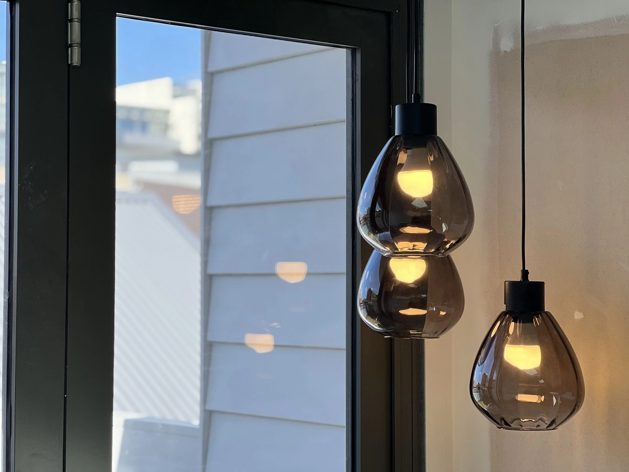 Close-up view of a modern pendant light with a glass shade, illuminated, hanging in front of a window with exterior siding visible outside.
