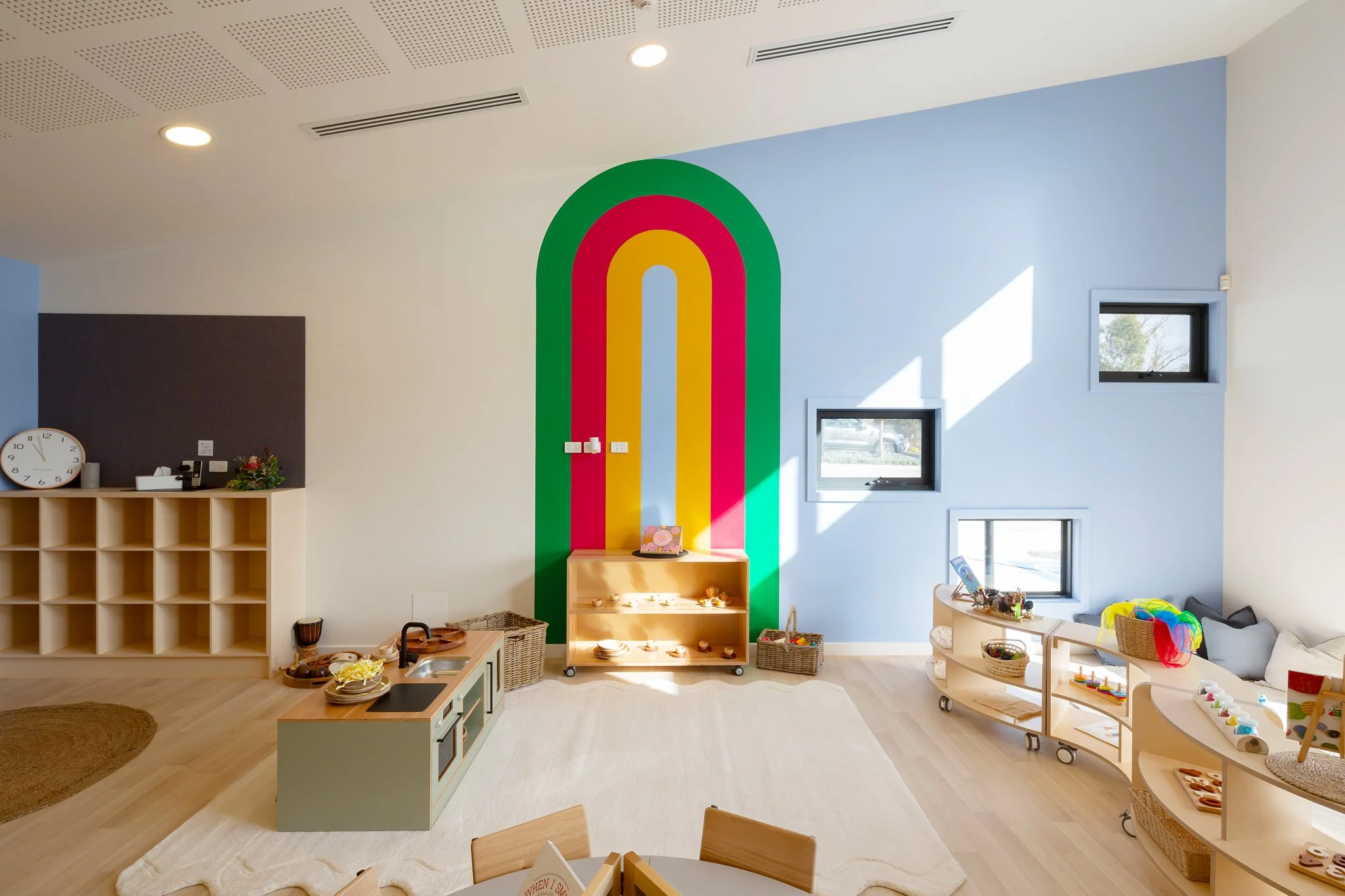 Bright playroom with a rainbow mural on the blue wall, shelves of toys, windows letting in sunlight, and a clock on the wall.