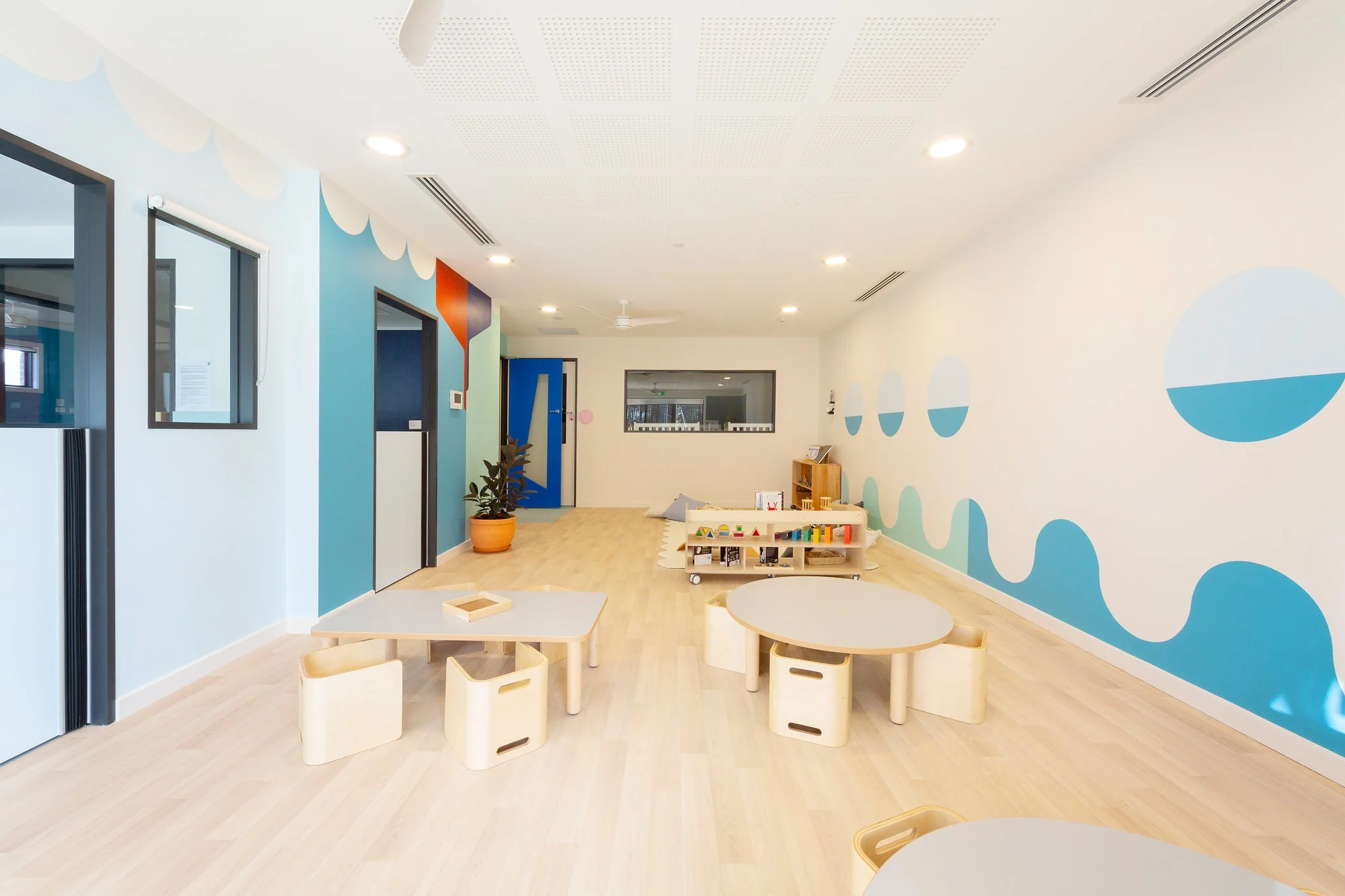 A brightly lit, modern classroom or activity room with light wood flooring and colorful wall murals featuring circles and abstract shapes. Small tables and chairs are arranged around the room, with a toy shelf and a potted plant near the door.